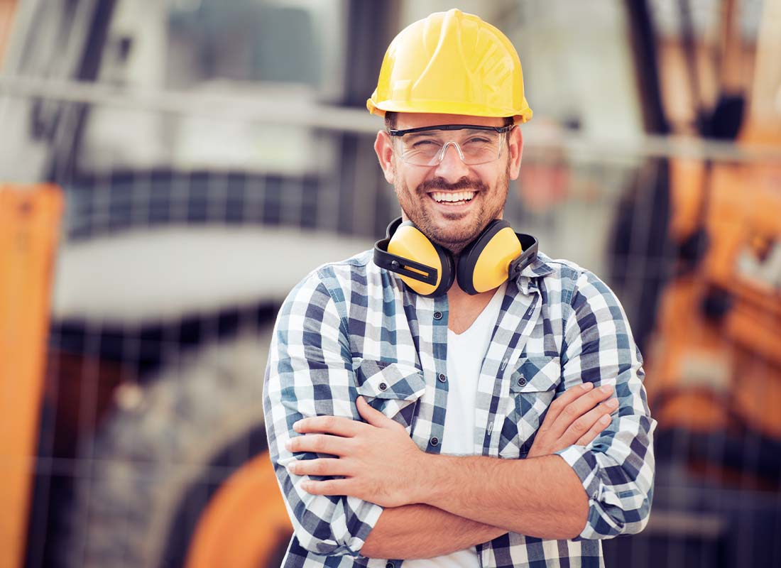 Contractor Insurance - Portrait of a Smiling Young Construction Worker in a Hard Hat with Arms Crossed Standing in Front Construction Equipment at a Construction Site
